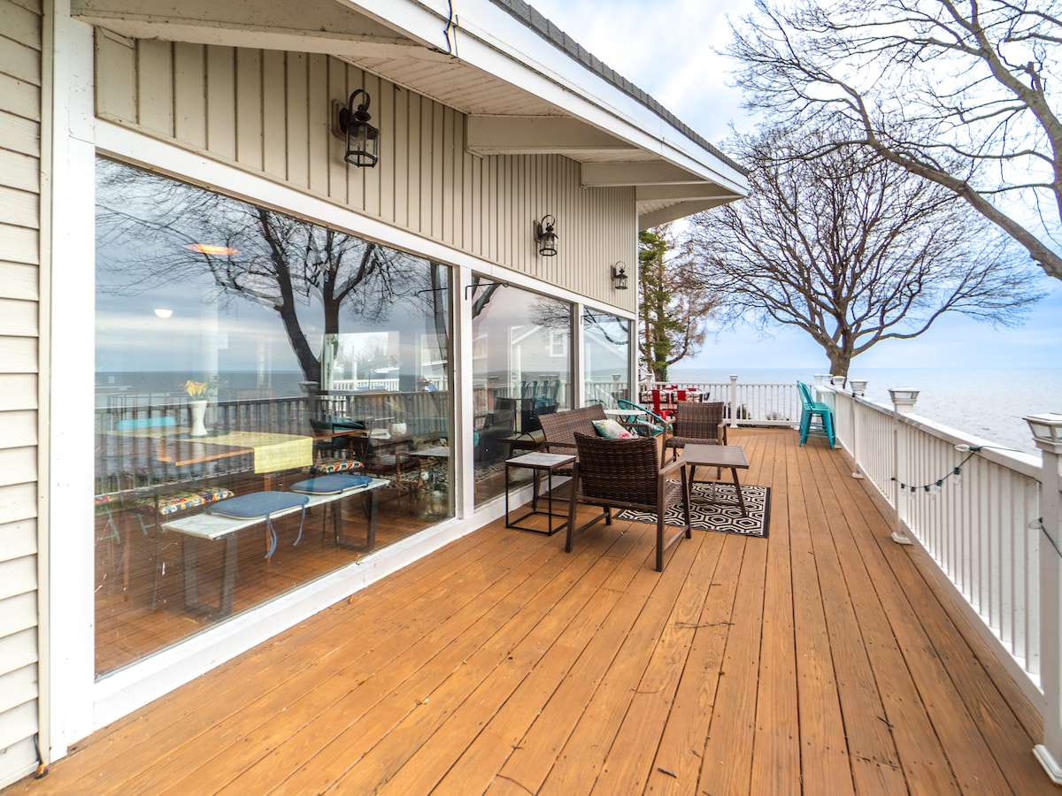 A spacious wooden deck extends over Lake Ontario, with large glass panels providing an unobstructed view of the water. Multiple seating areas are arranged, including woven chairs and a small table, surrounded by trees that gently frame the outdoor space.