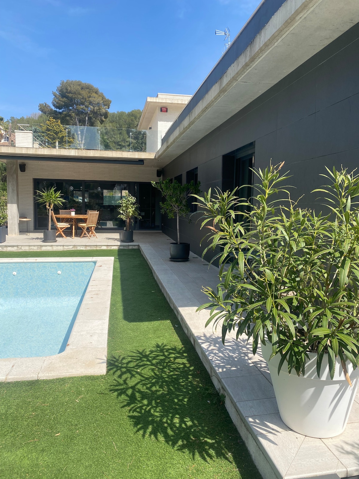 A modern home is depicted with a sleek exterior featuring large windows. A sparkling pool sits adjacent to a well-maintained garden area with potted plants. A small dining table made of wood is visible in the background under a shaded area.