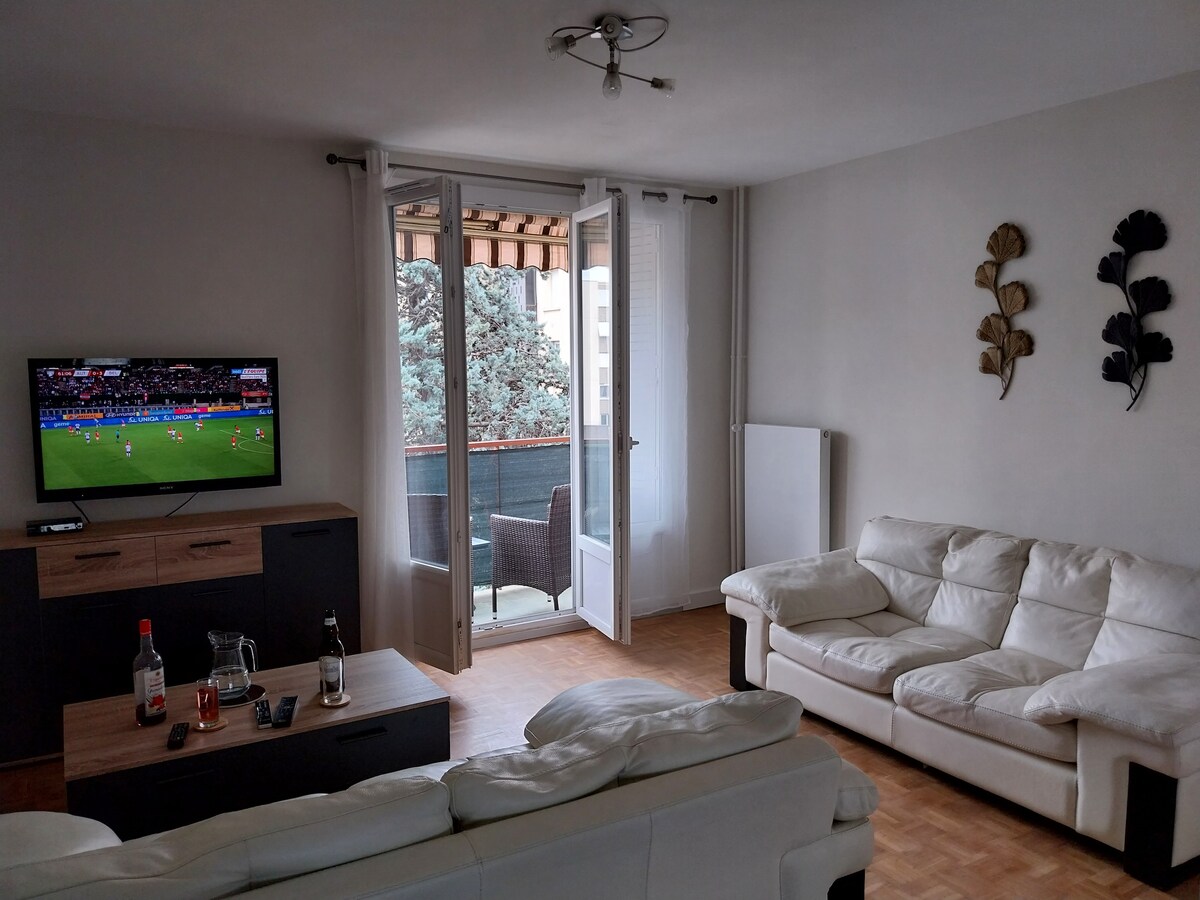 A spacious living area is furnished with a white sectional sofa and a wooden coffee table, which holds decorative items. A flat-screen television is mounted on the wall, displaying a sports event. Glass doors lead to a balcony with outdoor seating.