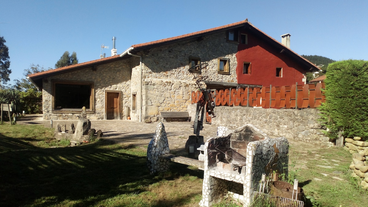 A charming stone house with a red accent wall is surrounded by a spacious outdoor area. A unique outdoor sculpture is visible in the foreground, while a well-kept lawn and wooden fencing contribute to the inviting surroundings.