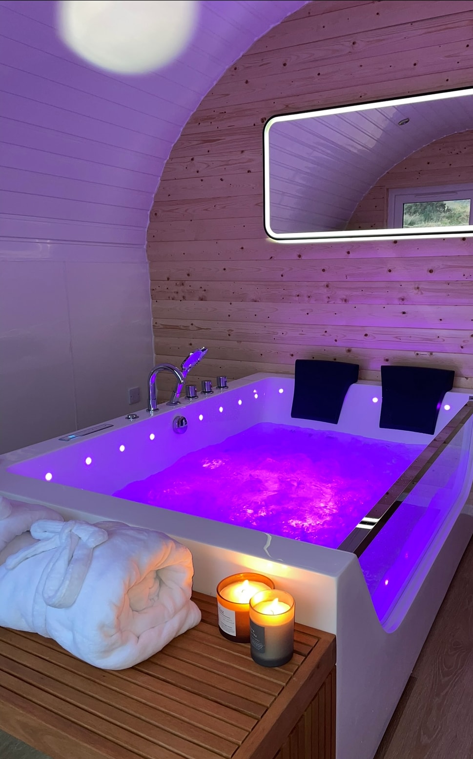 A jacquzzi bath pod is highlighted with soft purple lighting, creating a soothing ambiance. Two black towels are neatly placed at the edge, and a wooden surface holds two candles, providing additional warmth to the setting. The curved wooden walls enhance the intimate feel of the space.