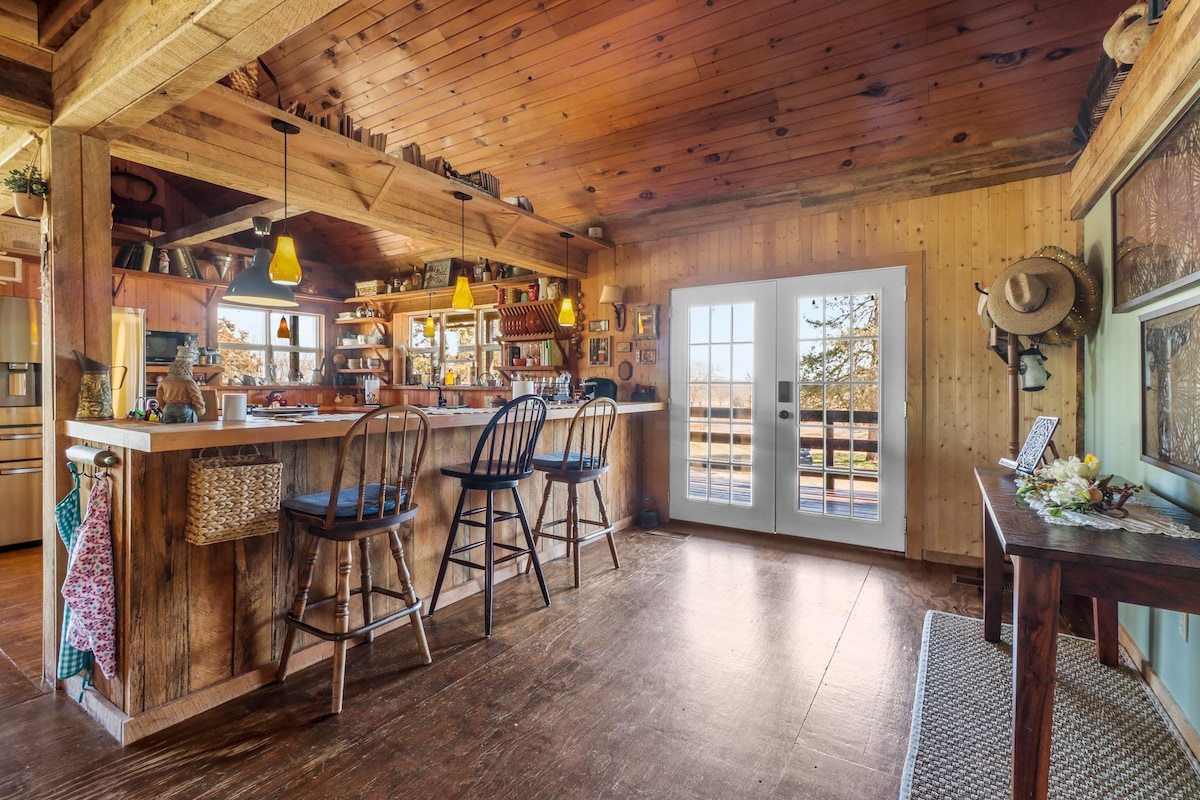 A warm and inviting kitchen area features wooden paneling and beams, with a bar equipped with three high-backed stools. Shelves display various kitchen items, and light streams through French doors that open onto the porch, offering a glimpse of the outdoor scenery.