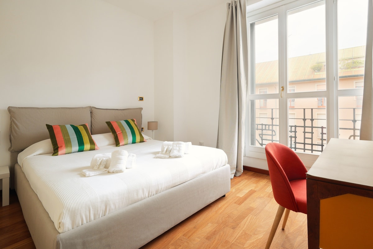 A bright bedroom features a spacious bed adorned with striped pillows. Natural light enters through large windows, illuminating the wooden floor. A small desk with a red chair is positioned against one wall, offering a comfortable work space.