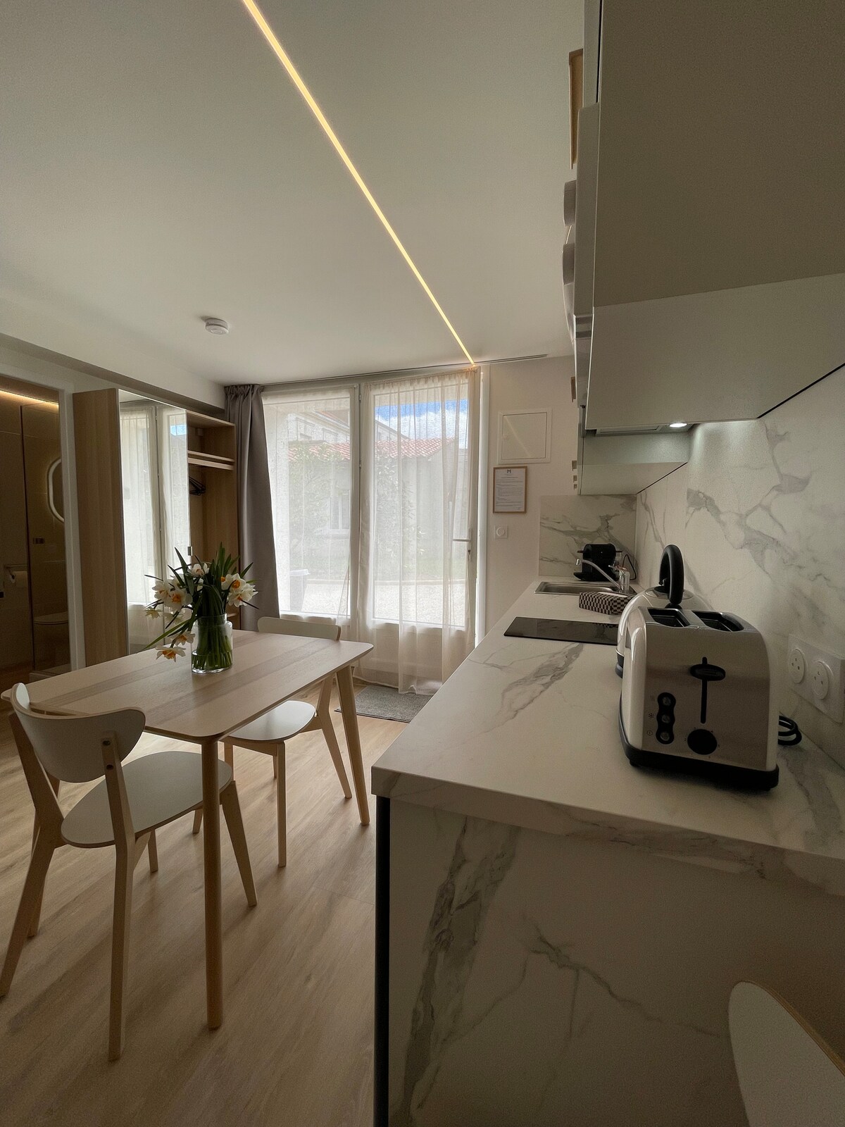 A modern kitchenette and dining area is displayed, featuring a sleek countertop with a toaster and a vase of fresh flowers. A wooden table with two chairs is positioned nearby, while large windows allow natural light to flood the space.