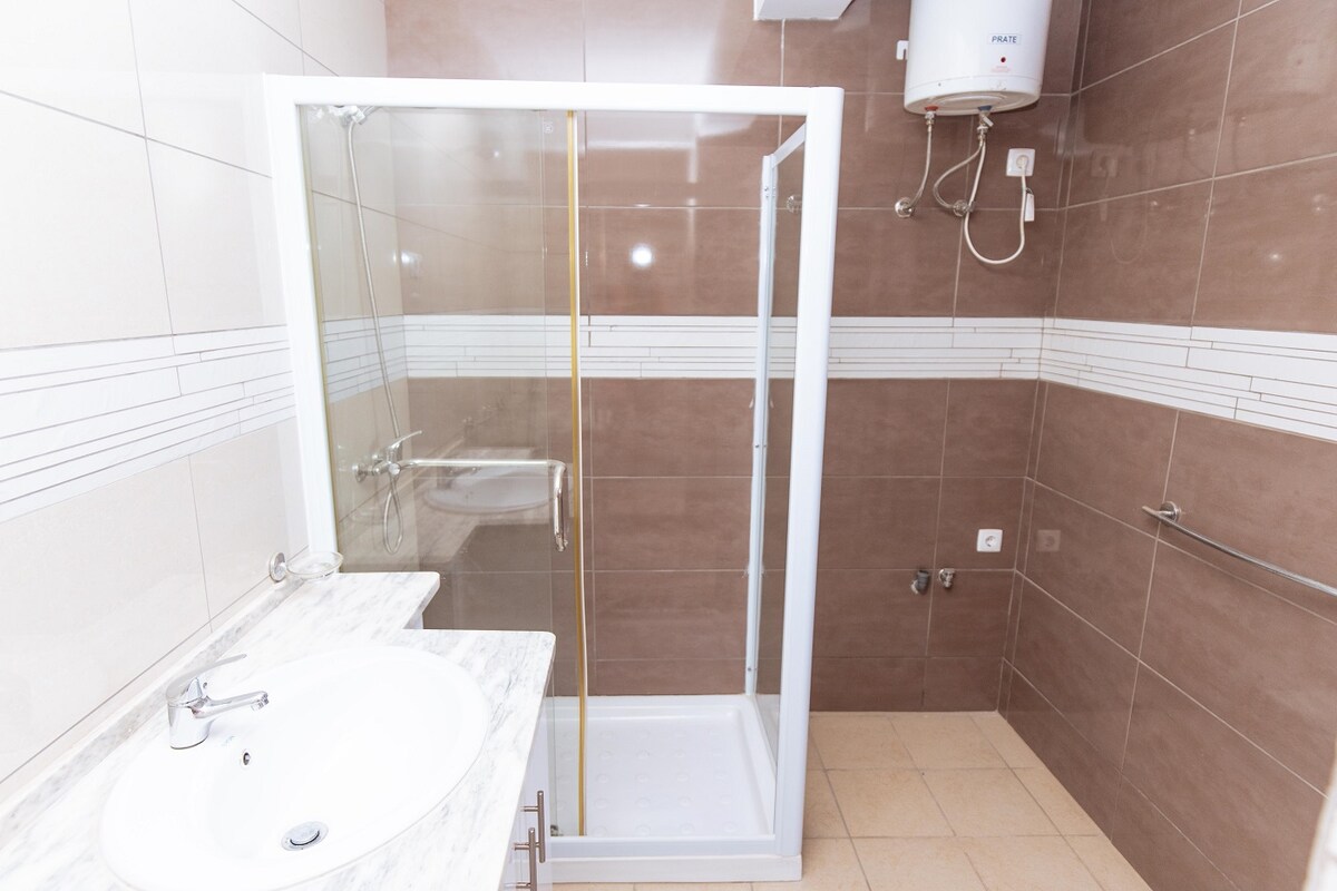 A modern bathroom features a shower enclosed in glass, with a white sink and a marble countertop. Neutral-toned tiles cover the walls, highlighted by simple horizontal lines. A towel rail and water heater are also visible, providing functional amenities.