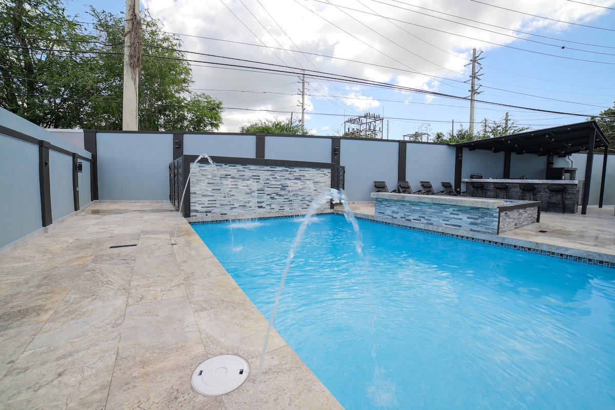 The pool area features a clear blue swimming pool with elegant fountains positioned along the edge. Surrounding the pool, a spacious deck is complemented by a lounge area with comfortable seating. A grill area is visible, with a covered terrace in the background.