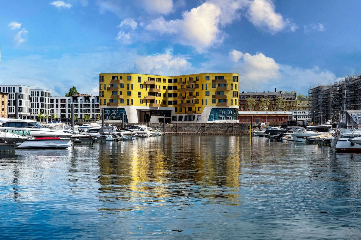 A modern waterfront building stands prominently along the harbor, featuring a striking yellow façade. The building reflects on the calm water, surrounded by various boats docked nearby. The scene captures a harmonious blend of contemporary architecture and serene water views.