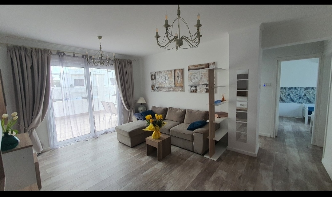 A living area features a comfortable sofa arranged around a central coffee table. Natural light enters through large glass doors leading to a veranda. Neutral-toned curtains frame the windows, enhancing the fresh decor. A cluster of decorative flowers sits on the table, adding a touch of color.