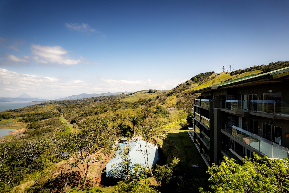 A scenic view captures the expansive landscape of rolling hills and lush greenery, with a serene lake visible in the distance. The modern building features multiple balconies, inviting guests to enjoy the tranquil surroundings while being embraced by nature.