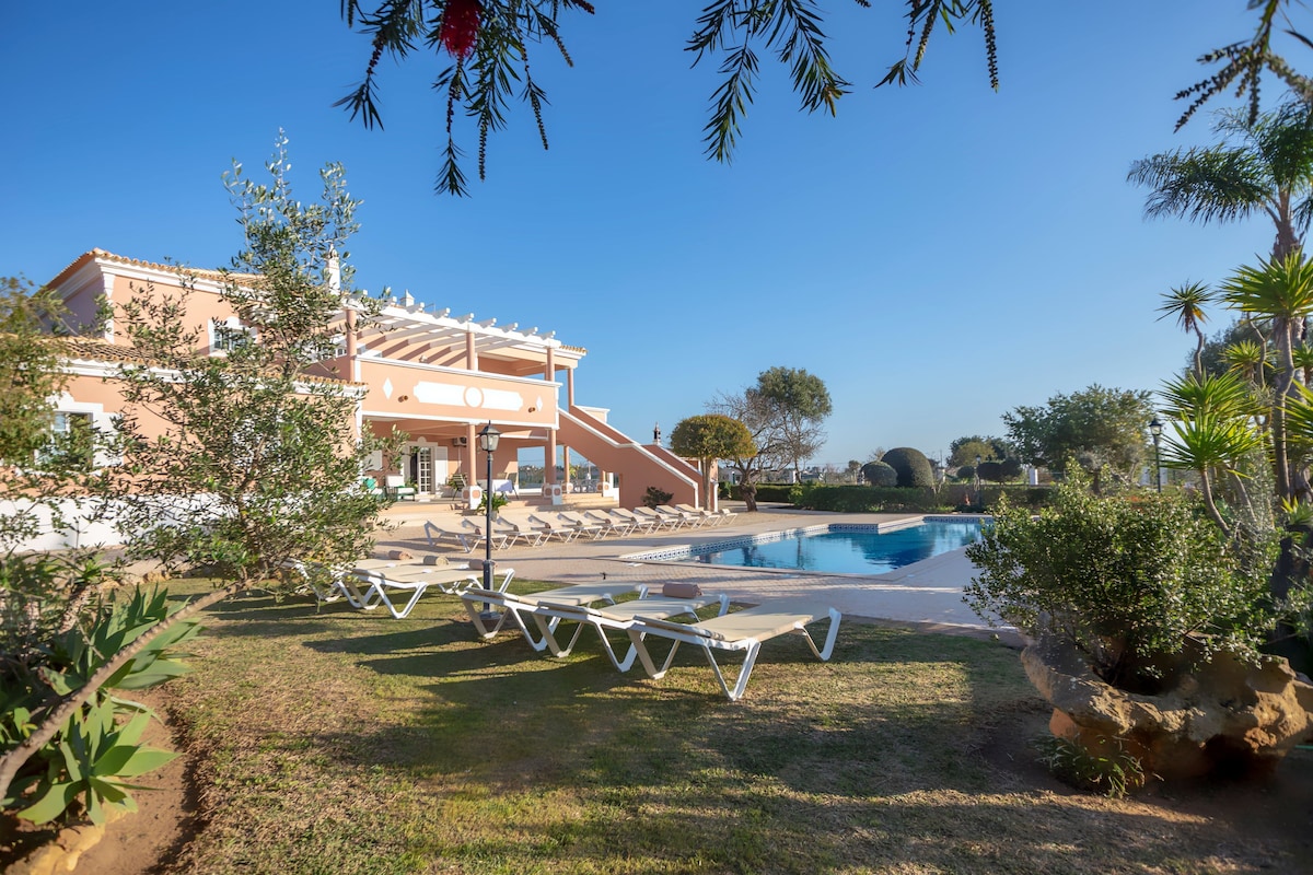 The exterior of the guesthouse is displayed, showcasing a two-story structure with light-colored walls and a spacious terrace. Lounge chairs are arranged around the outdoor pool, surrounded by well-maintained greenery. Clear skies and natural landscaping contribute to a serene outdoor environment.