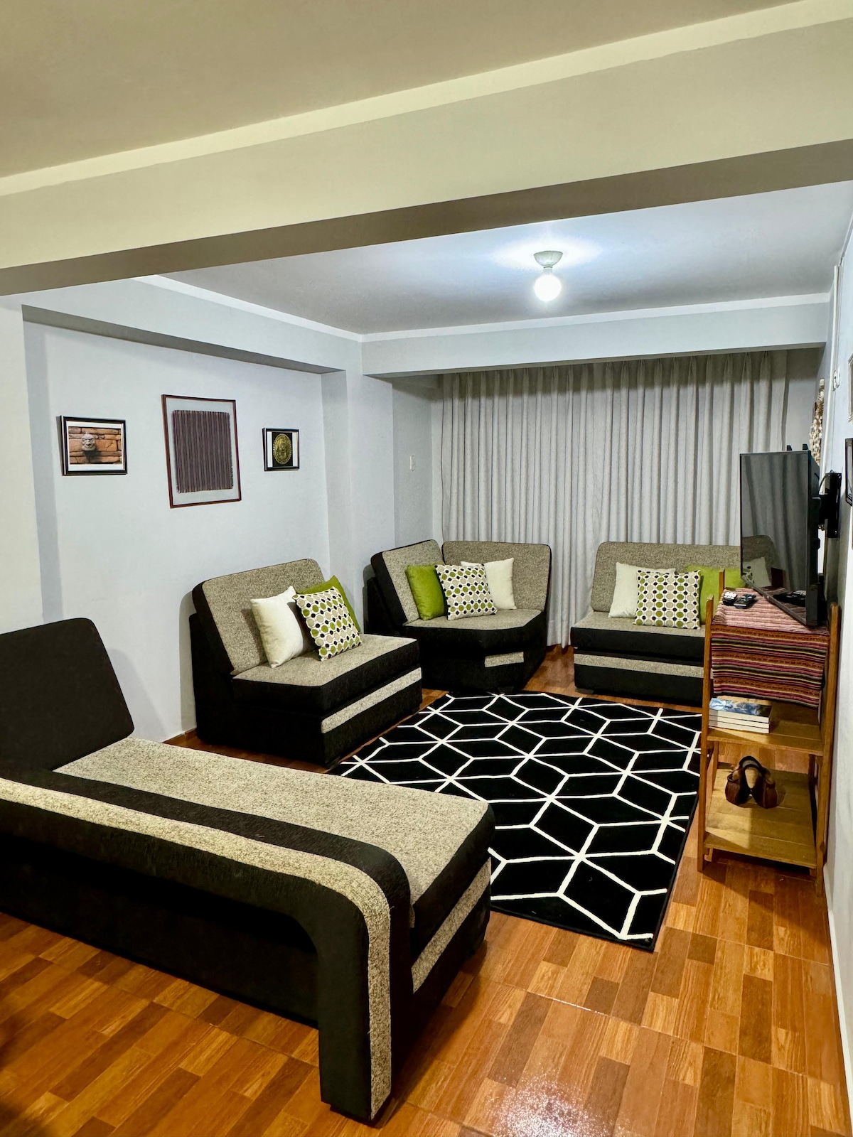 A comfortable living area is equipped with four modern armchairs adorned with decorative cushions. A geometric-patterned rug covers the hardwood floor, and light enters through a large window with sheer curtains. Minimal artwork is displayed on the walls, adding a touch of style.
