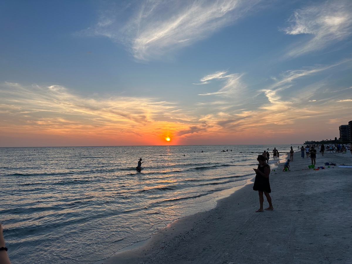 A serene beach scene captures the setting sun on the horizon, casting warm orange and pink hues across the sky. Gentle waves lap against the shore, while beachgoers stroll along the sand, some enjoying the water, creating a peaceful atmosphere.