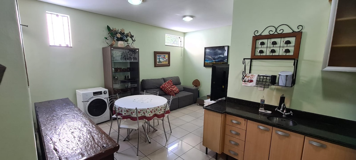 A well-lit living area is displayed with a gray couch decorated with cushions. A round dining table sits in the center, surrounded by chairs. A glass display cabinet and a washing machine are present, along with a kitchen area featuring wooden cabinetry and a dark countertop.