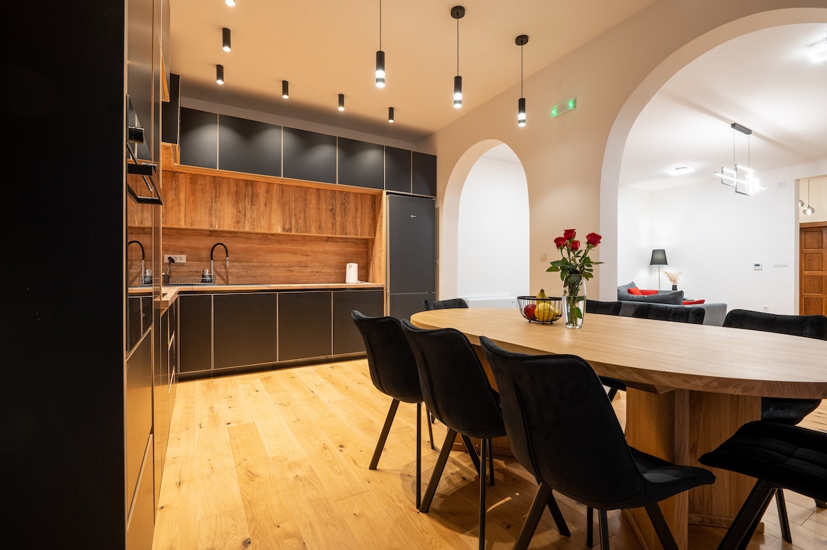 A modern kitchen showcases sleek black cabinetry paired with warm wooden accents. A spacious round dining table is surrounded by black chairs, while soft lighting illuminates the area. Fresh flowers are displayed on the table, adding a touch of color to the design.