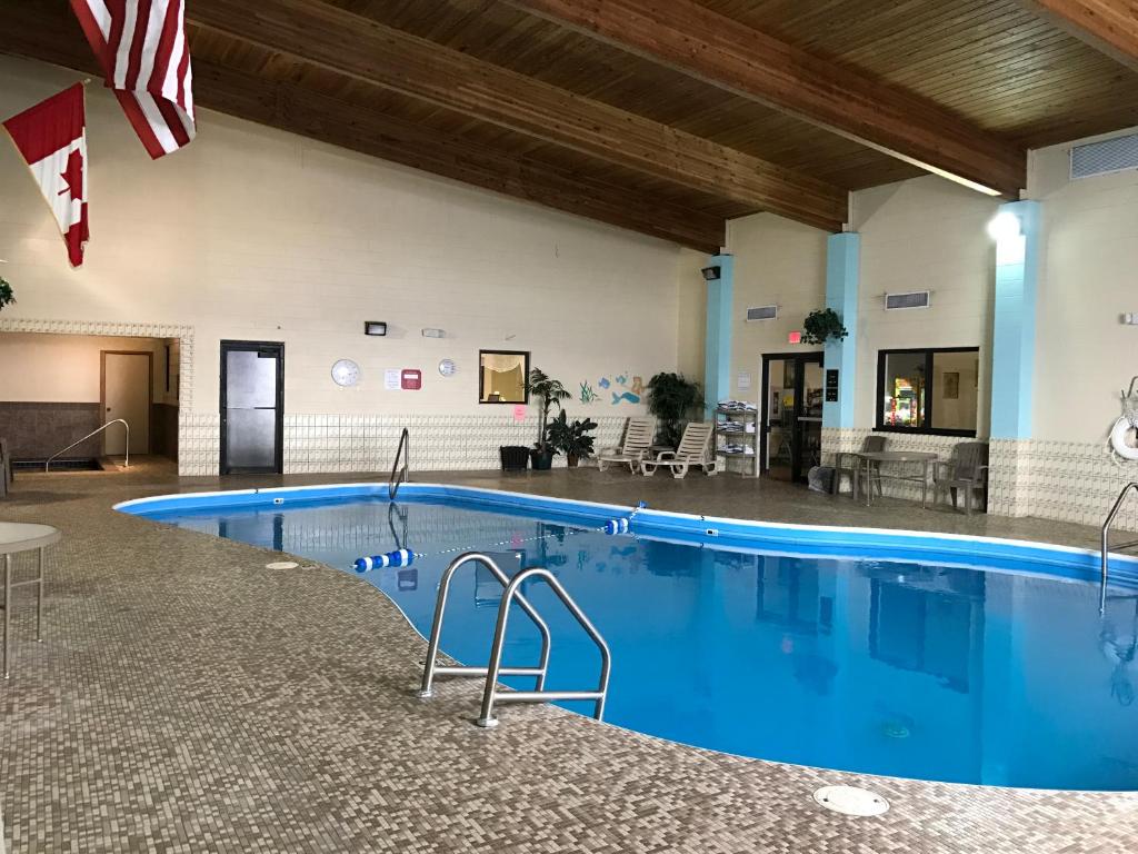 An indoor swimming pool is featured, surrounded by a mosaic tiled floor. Flags hang from the ceiling, while lounge chairs are positioned nearby. Warm wooden beams enhance the overhead space, contributing to a bright and inviting atmosphere.