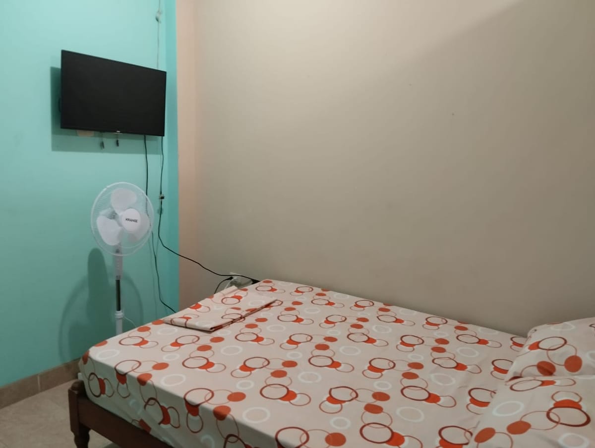 A simple sleeping area features a spacious bed with a patterned duvet cover. A small television is mounted on the wall, with a fan positioned nearby. The walls are painted in soft colors, contributing to a calm environment.