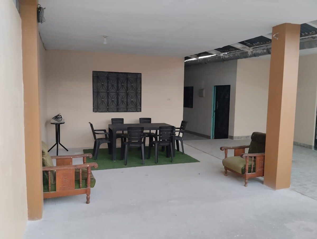 An outdoor seating area is featured, consisting of a large dining table surrounded by multiple chairs and a small side table. Comfortable armchairs provide additional seating. The space is partially covered, with a green carpet covering the floor and a window allowing natural light to enter.