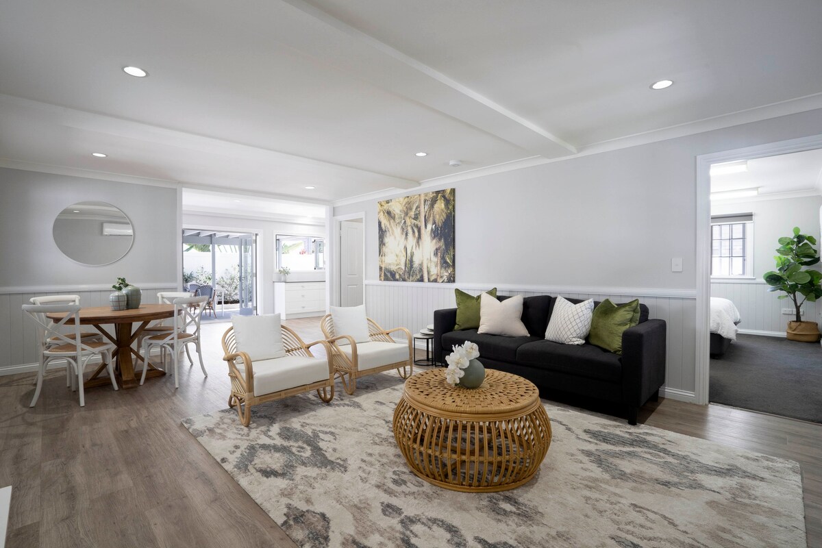 A bright and inviting living area features comfortable seating arranged around a decorative coffee table. A dining table for four sits nearby, with soft natural light flowing in from the adjoining space. The aesthetic is complemented by modern decor and neutral tones.
