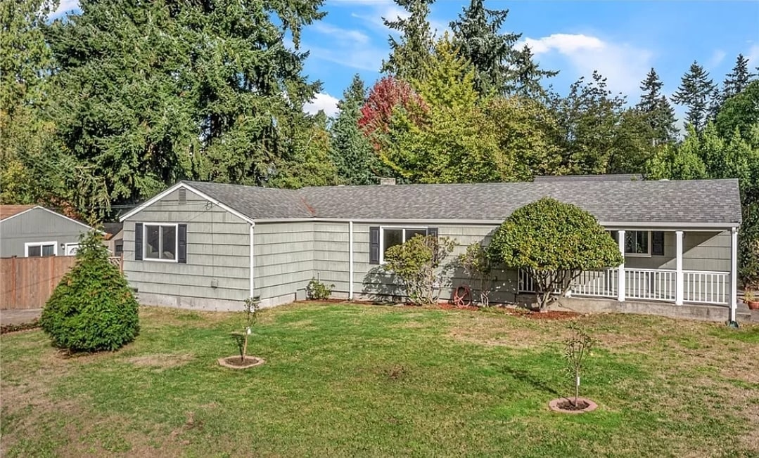 A single-family home is showcased with a light gray exterior and a manicured yard featuring circular flower beds. The surrounding greenery includes trees in various shades, contributing to a natural setting. A covered porch is visible at the front of the house.