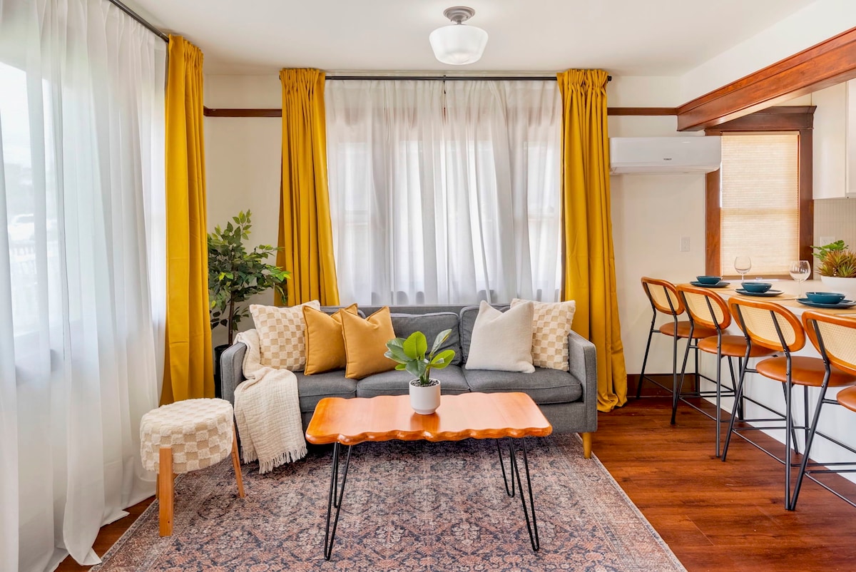 The living room features a comfortable gray sofa adorned with patterned cushions and a cozy throw. A wooden coffee table is positioned in front, complemented by a small plant. Bright yellow curtains frame the windows, allowing natural light to fill the space.