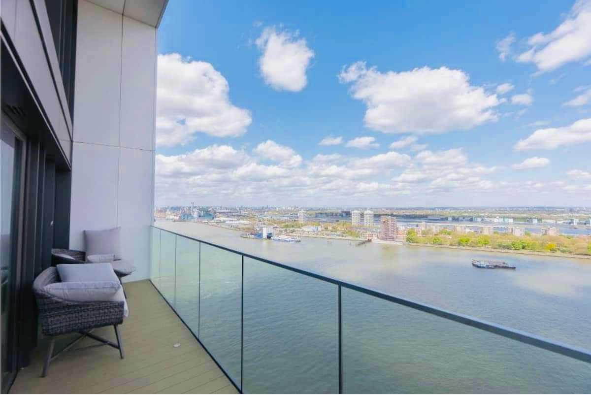 A private balcony overlooks the River Thames, showcasing panoramic views of the water and cityscape. Two comfortable chairs are placed against the glass railing, allowing for unobstructed sightlines of the horizon and sky, which is dotted with fluffy clouds.