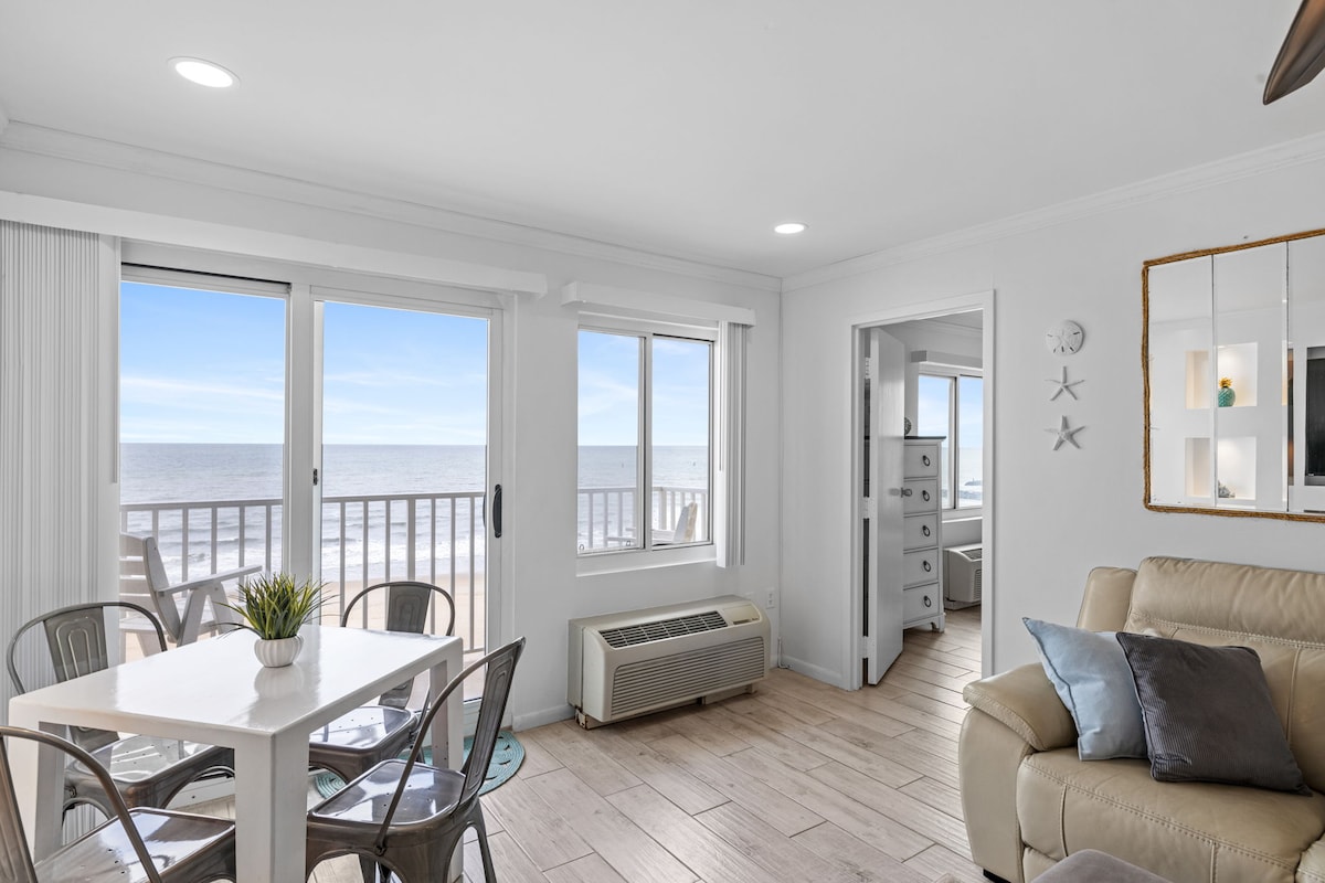 A bright living area features large windows with ocean views, allowing natural light to fill the space. A dining table for four is positioned beside the window, while a comfortable beige sofa is arranged to create a welcoming atmosphere.