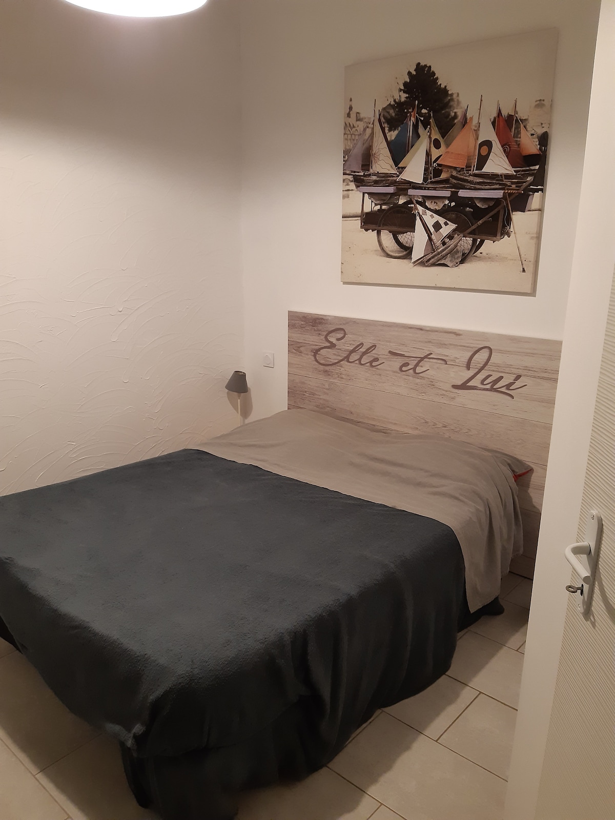 A cozy sleeping area features a spacious bed with a gray blanket. Above the bed, a framed image of sailboats adds a decorative touch to the room. A bedside lamp is mounted on the wall, and light-colored tiles cover the floor.