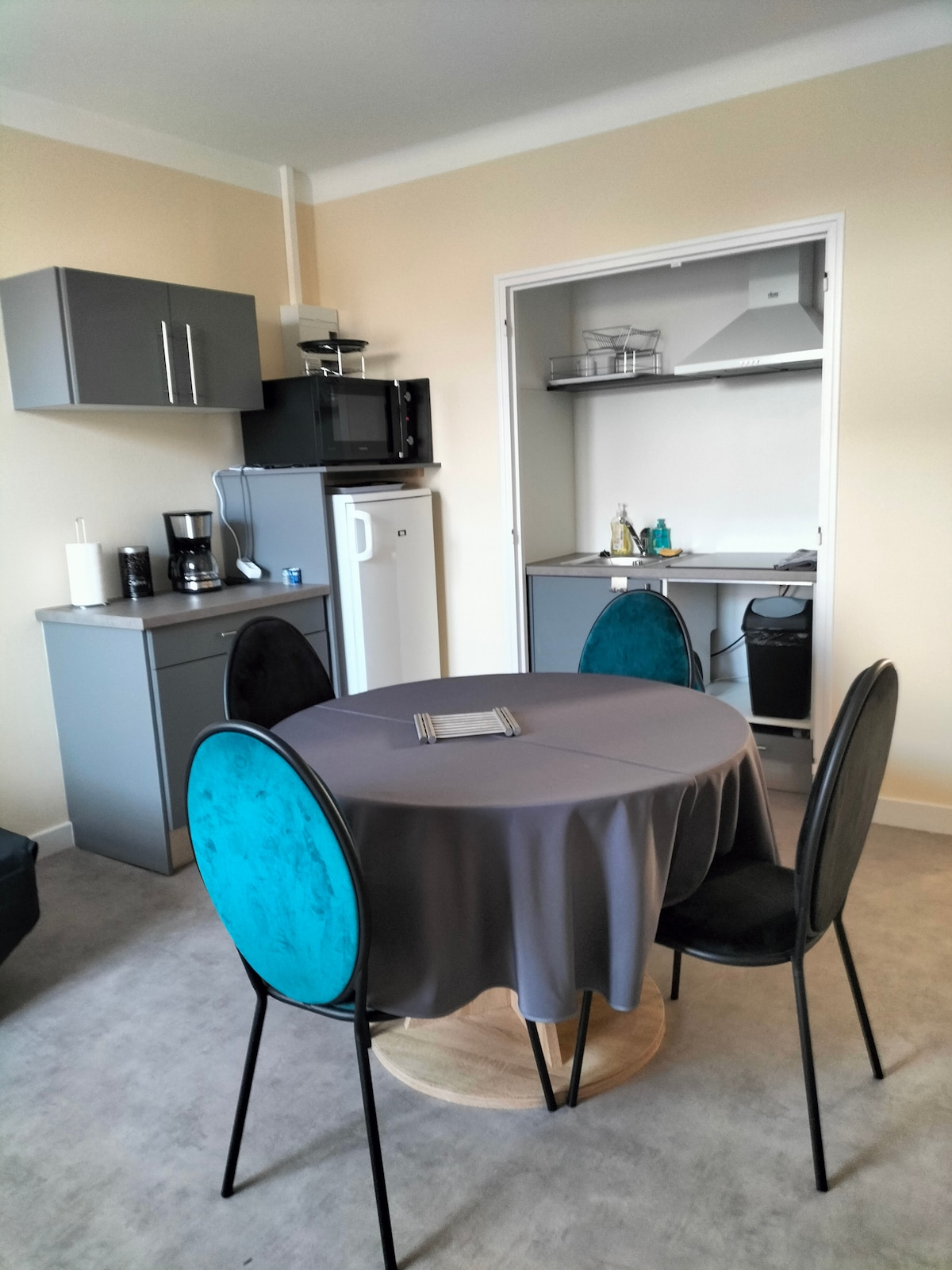 A modern kitchen area features grey cabinetry paired with a round dining table surrounded by four black and teal chairs. Appliances visible include a microwave, refrigerator, and coffee maker. A small shelving unit is positioned in the corner, contributing to the functional layout.