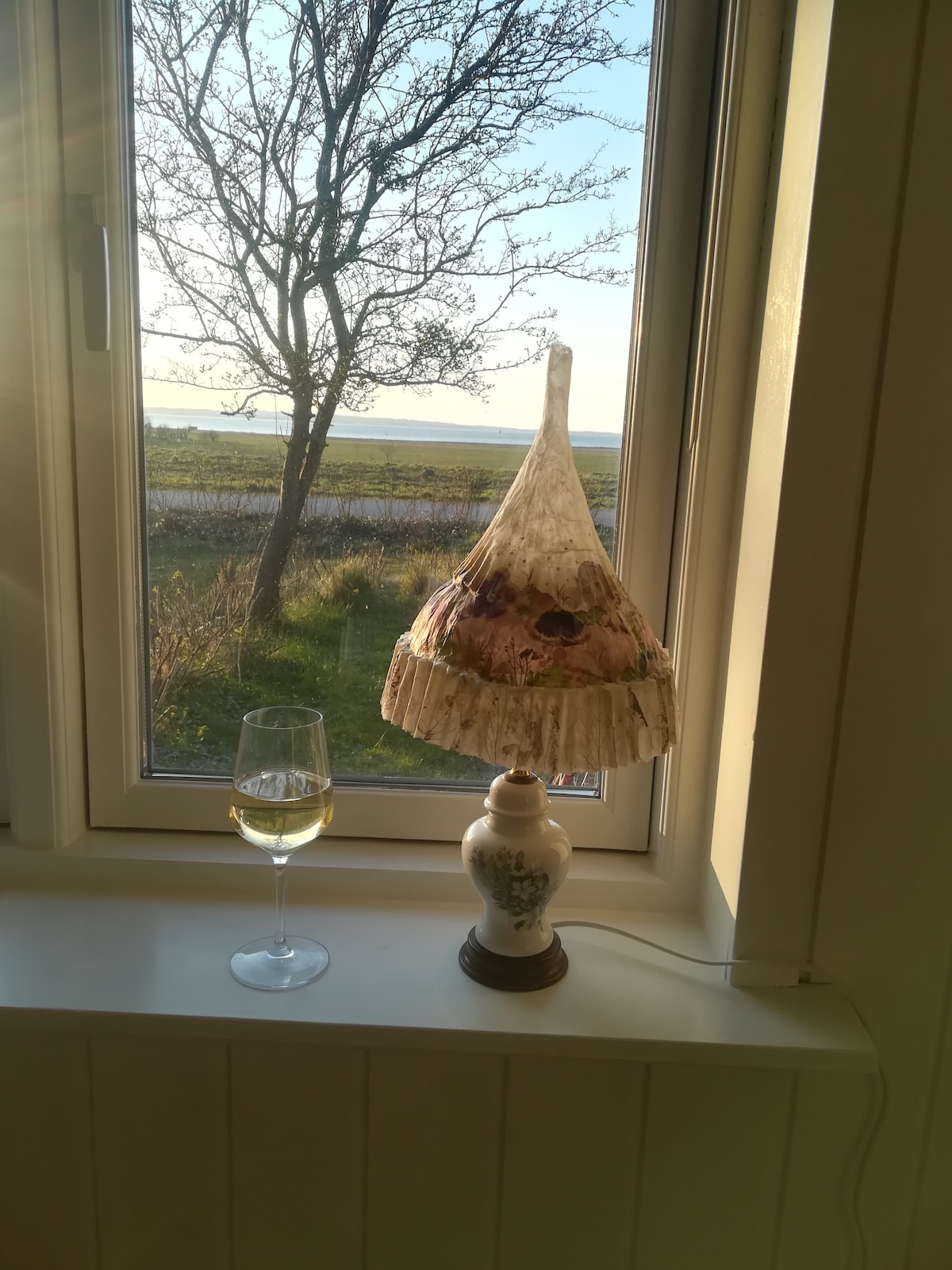 A decorative lamp with a floral shade is positioned on a windowsill, casting soft light. Next to it, a glass of white wine reflects the sunlight. A scenic view of greenery and a distant horizon is visible through the window, enhancing the serene environment.