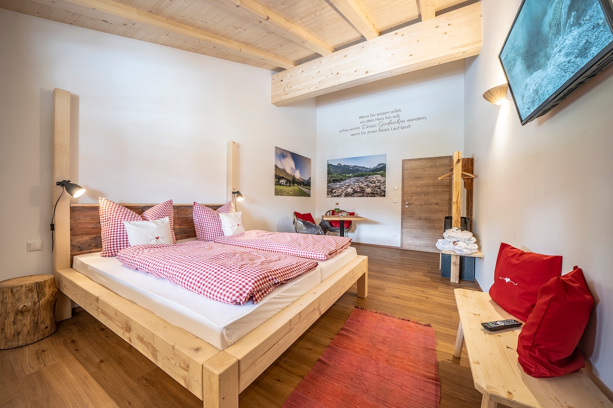 A spacious room features two wooden beds adorned with red and white checkered bedding. Cozy seating options are positioned against the wall, with artwork depicting scenic landscapes. Natural wood accents and warm lighting create a welcoming environment, complemented by a flat-screen TV mounted on the wall.