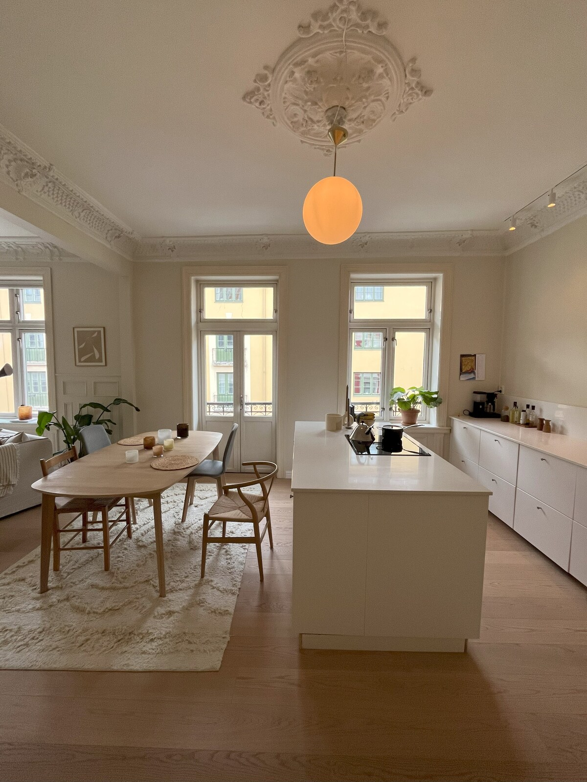 A modern kitchen and dining area is featured, characterized by a spacious dining table surrounded by wooden chairs. Natural light streams through large windows, illuminating the white cabinetry and island. Simple decor elements, such as plants and tasteful lighting, add to the inviting ambiance.
