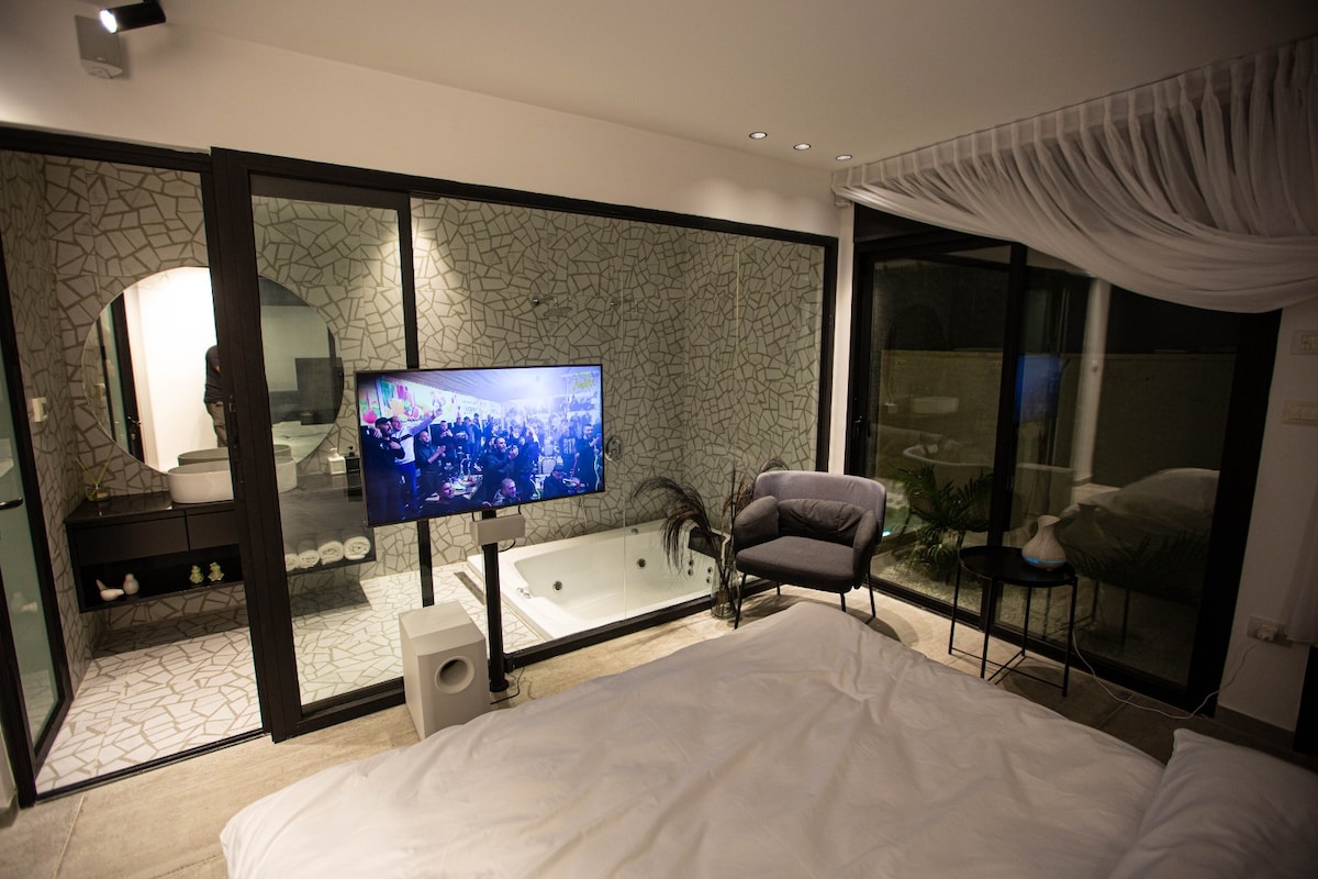 A modern bedroom is illuminated by soft lighting, featuring a spacious bed covered with white linens. A sleek television is positioned in front of the bed, and large glass walls provide a view of a stylish bathroom area with a stone design. An armchair is situated beside a small table.