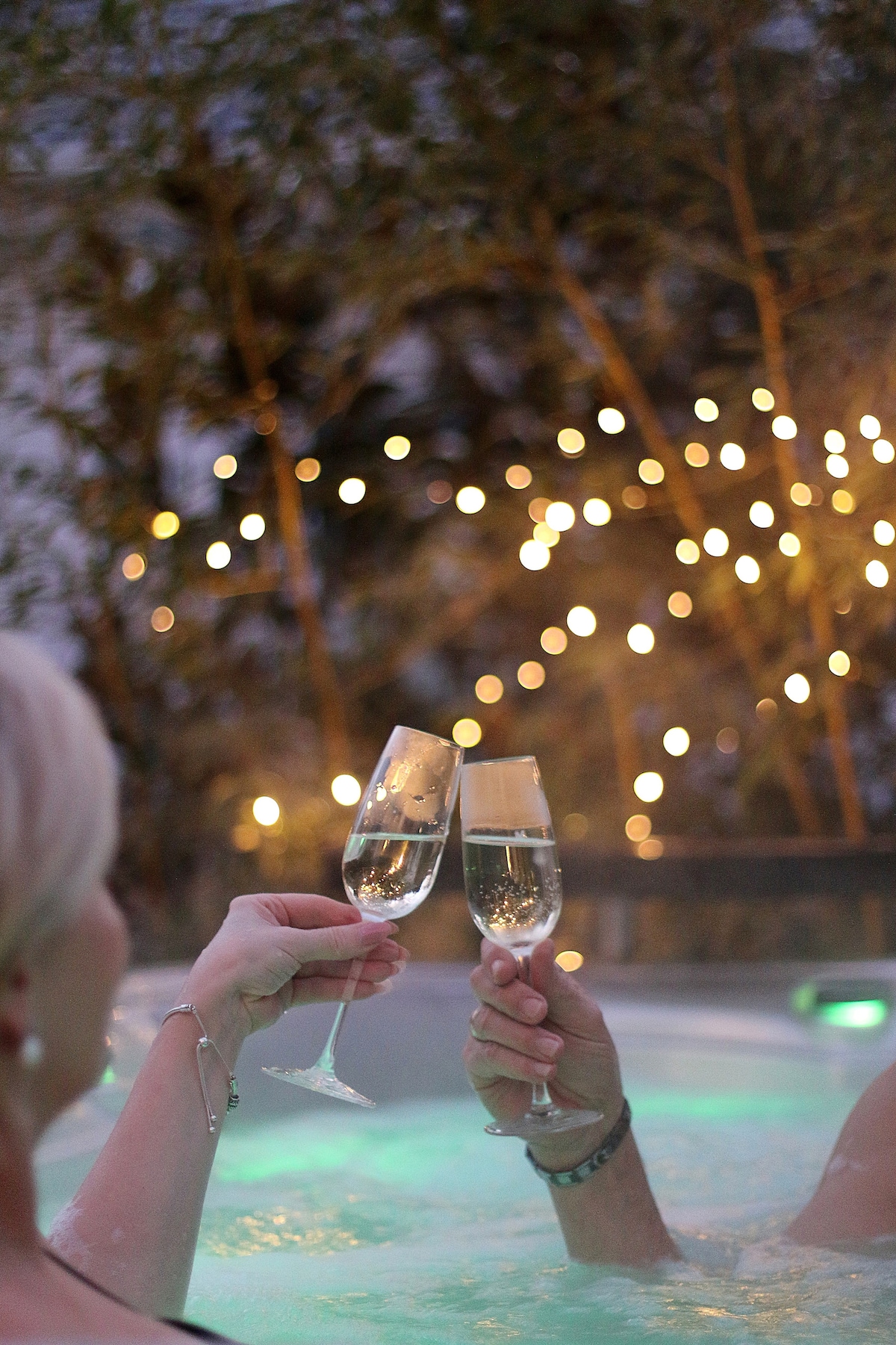 Two champagne flutes are raised in a toast, with light bokeh creating a soft, glowing backdrop. The scene is set in a spa-like environment, hinting at relaxation and enjoyment. The warm light enhances the atmosphere of celebration.