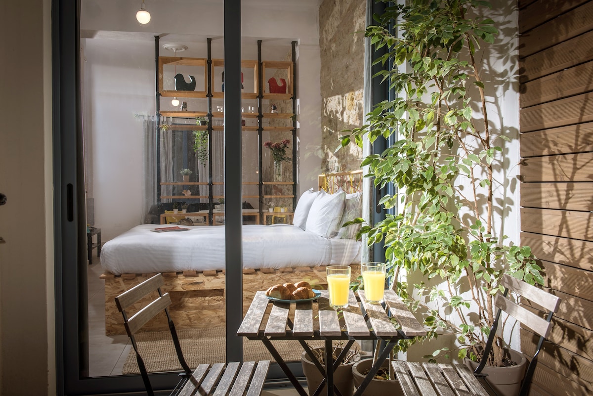 A small outdoor seating area is highlighted by a wooden table and two chairs, set for breakfast with fresh croissants and two glasses of orange juice. Glass doors reveal a bright bedroom beyond, adorned with a wooden pallet bed and greenery.