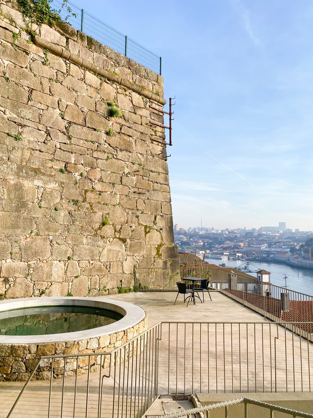 A spacious outdoor terrace features a round stone hot tub overlooking the river. A single black chair is positioned nearby, with the terrace's wooden flooring extending towards a scenic view of the river and cityscape beyond.