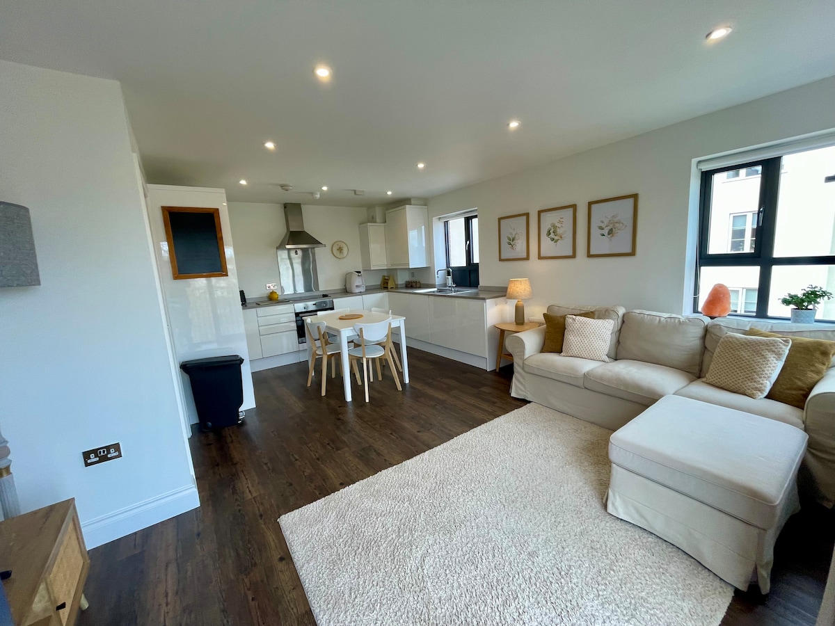A modern living space is presented with a comfortable sectional sofa and a soft area rug. A dining table with four chairs is positioned nearby, while the kitchen features sleek cabinets and a stainless steel stove. Natural light fills the room through large windows.