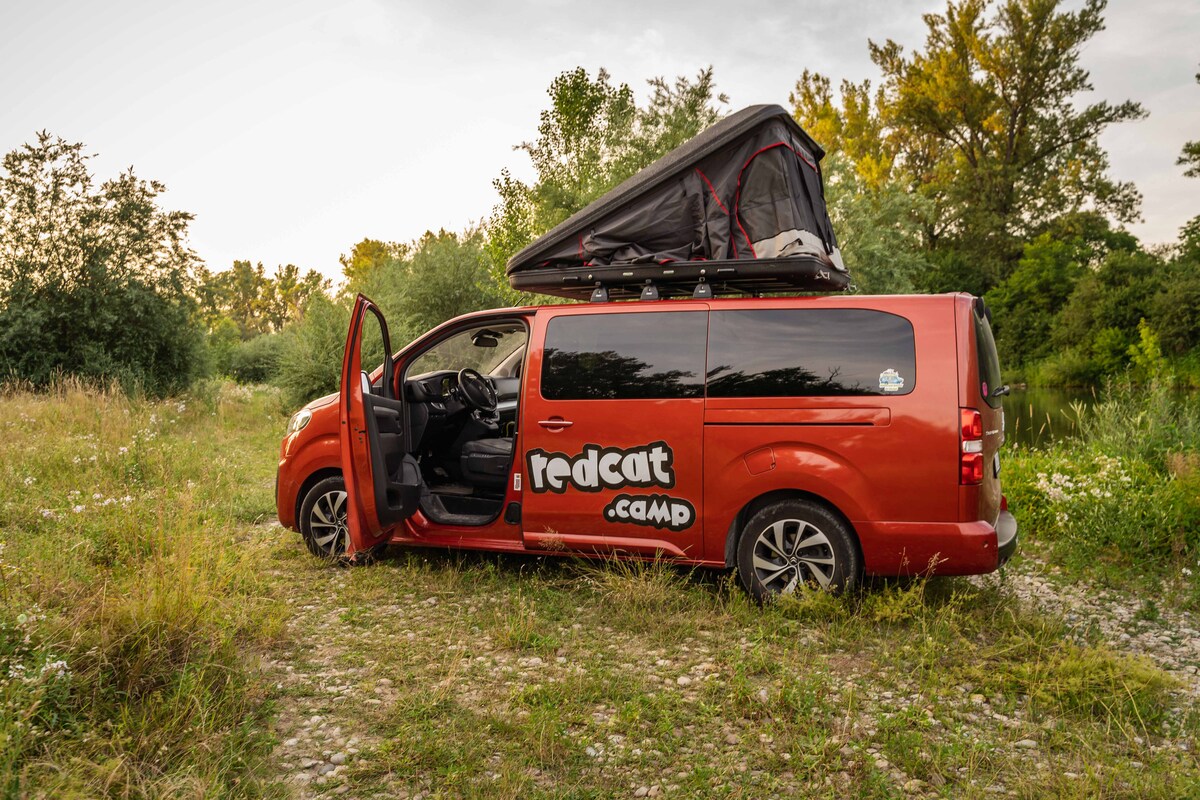 A red van is parked on a gravel surface, surrounded by greenery. The van features an opened pop-up roof tent, showcasing a compact sleeping area above. The driver's side door is ajar, providing a glimpse into the interior. Soft evening light casts a calming ambience.