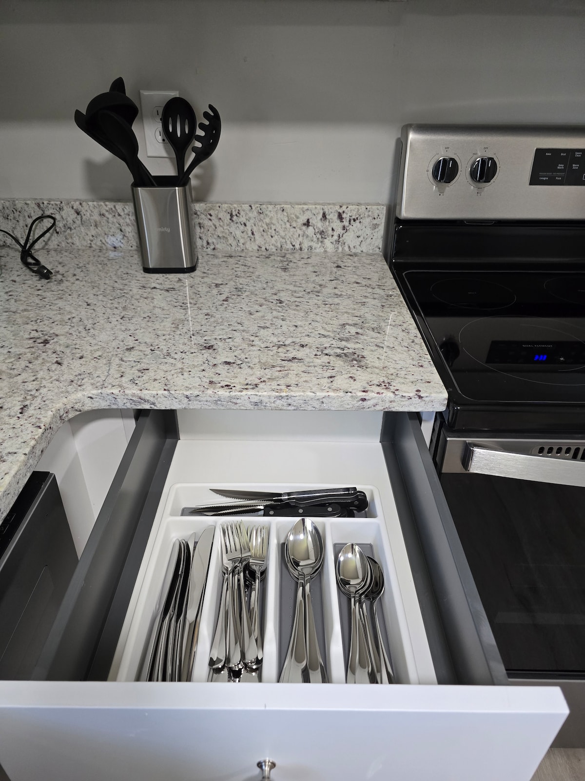 A well-organized kitchen drawer is displayed, featuring a clean cutlery tray filled with various utensils, including forks, knives, and spoons. A smooth granite countertop is visible above, with a stove and utensils holder nearby, emphasizing the functional kitchen space.