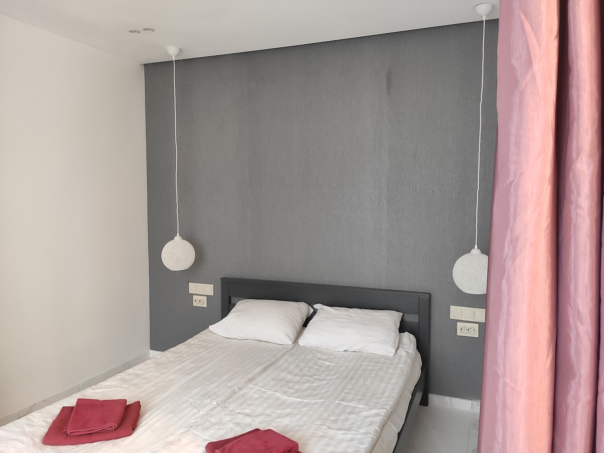 A minimalist bedroom is presented with a light-colored wall and a contrasting dark accent wall. The spacious bed features two white pillows, while red towels are neatly placed at the foot. Soft pendant lights hang above each side of the bed, enhancing the serene atmosphere.