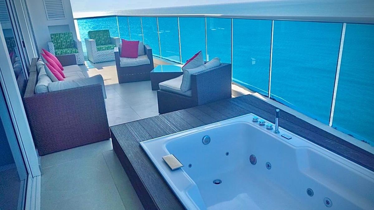 A spacious terrace is presented, featuring a private Jacuzzi and a comfortable seating area. The seating arrangement includes a large sofa, two plush armchairs, and a coffee table, all framed by glass railings that offer a clear view of the ocean.