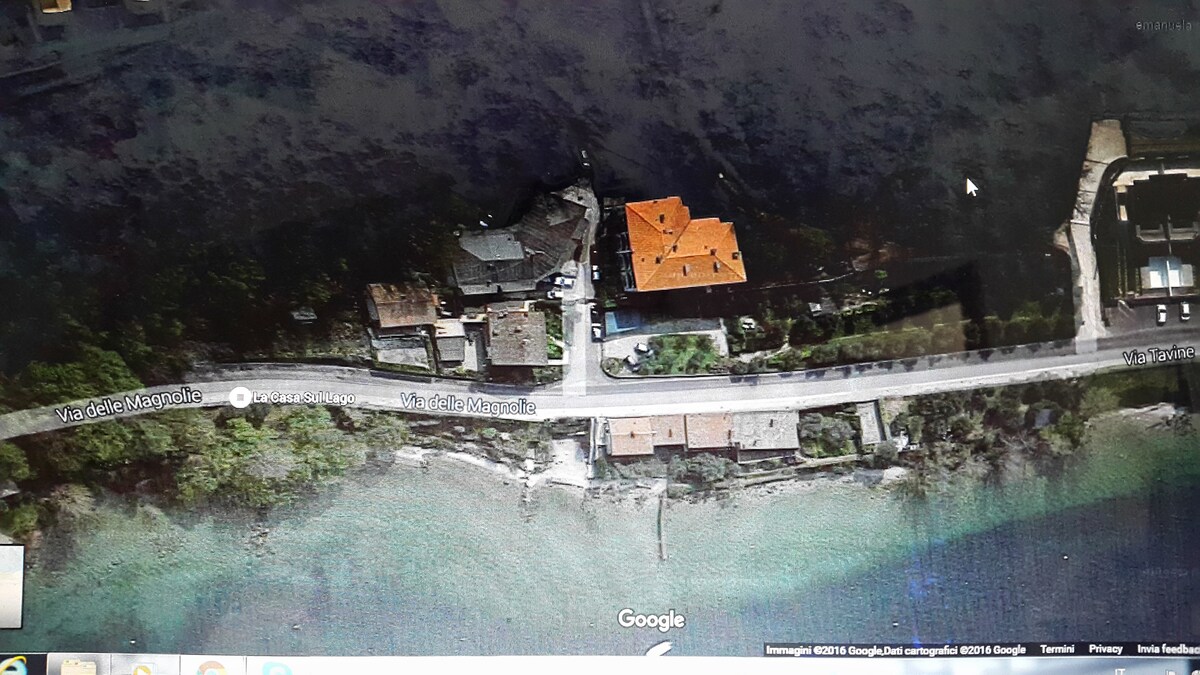 An aerial view showcases a serene lakeside area with a private road running alongside. Several structures are visible, including buildings with distinct roof styles. The surrounding greenery merges with the water, creating a rural, tranquil environment near a picturesque coastline.