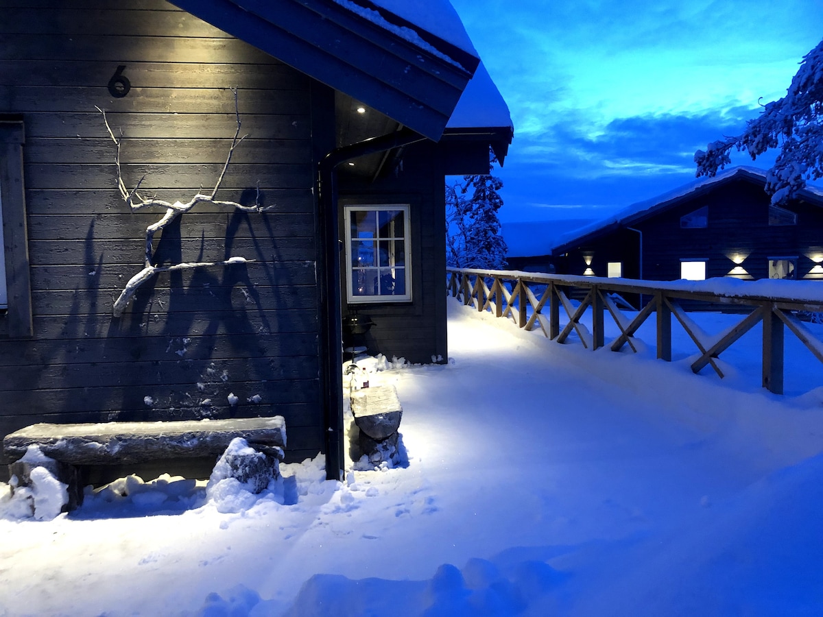 A wooden structure is depicted in a snowy landscape, illuminated by soft outdoor lighting. A bench sits under the covered area beside the entrance, while a railing extends along the pathway, leading towards additional buildings in the background. The evening sky casts a tranquil blue hue.