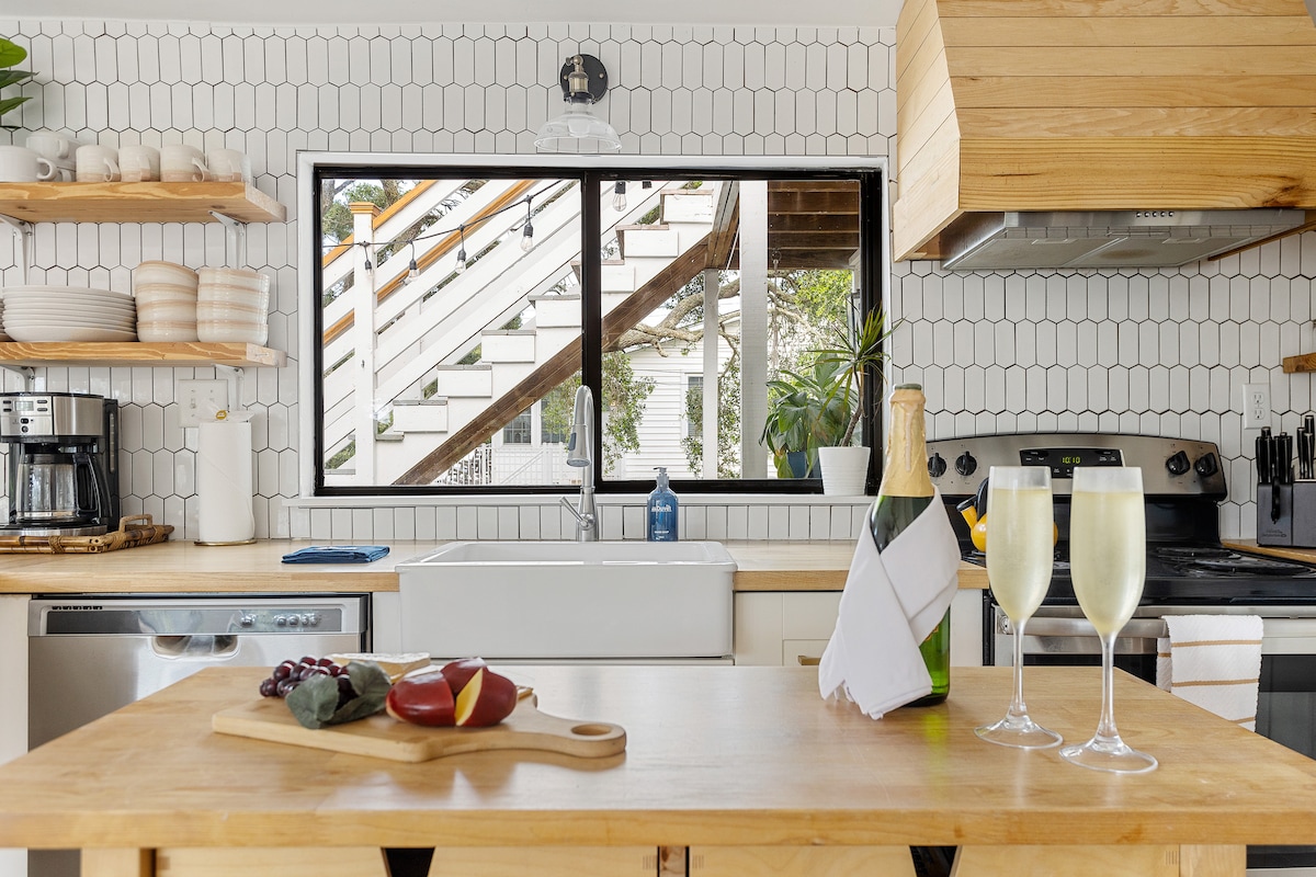 A fully equipped kitchen is featured, showcasing wooden countertops and white tiled walls. A farmhouse sink is positioned below a large window, allowing natural light to fill the space. Nearby, a charcuterie board displays sliced fruits, and two glasses of champagne are elegantly presented.