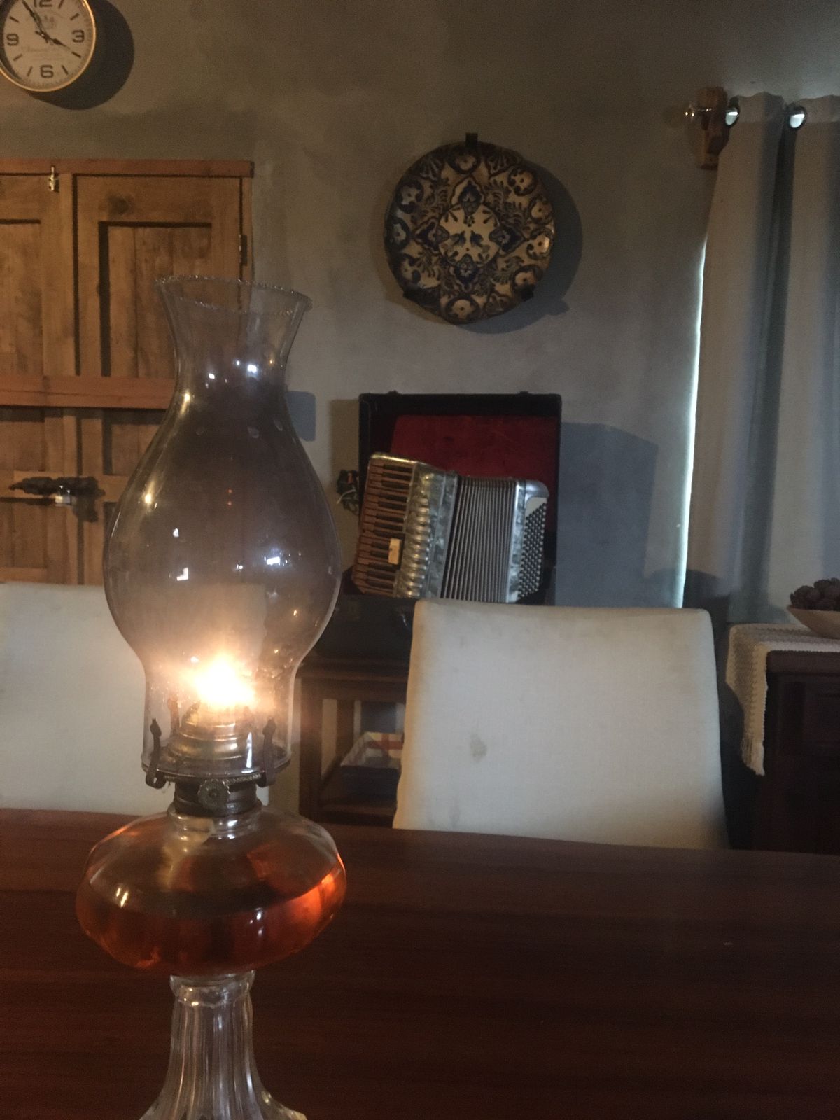 A rustic glass oil lamp illuminates a wooden table, casting a warm glow. In the background, a decorative wall clock and an ornate wall plate complement a vintage accordion, contributing to the cozy atmosphere of the interior space.