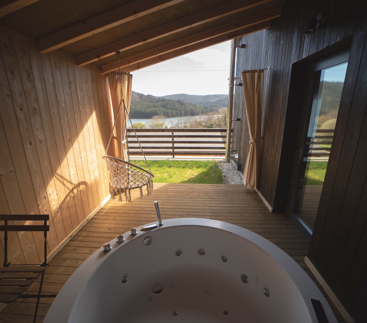 An outdoor terrace features a covered area with wooden walls and large glass doors, providing views of the surrounding mountains and greenery. A hot tub is positioned on the deck, while a hanging chair adds a cozy touch to the space.