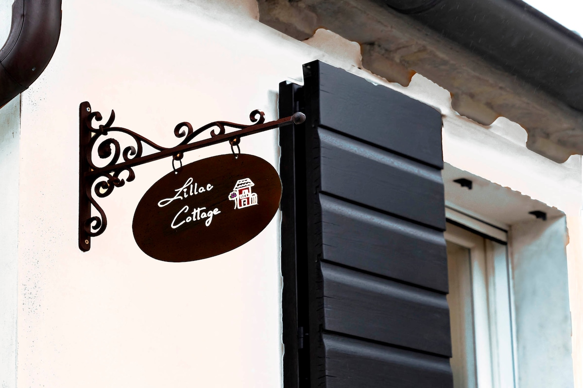 A decorative sign is mounted beside a window, showcasing the name 'Zillo Cottage' in an elegant font. The sign is supported by an ornate bracket, adding a touch of charm to the exterior. The window is framed with dark shutters, complementing the overall design.