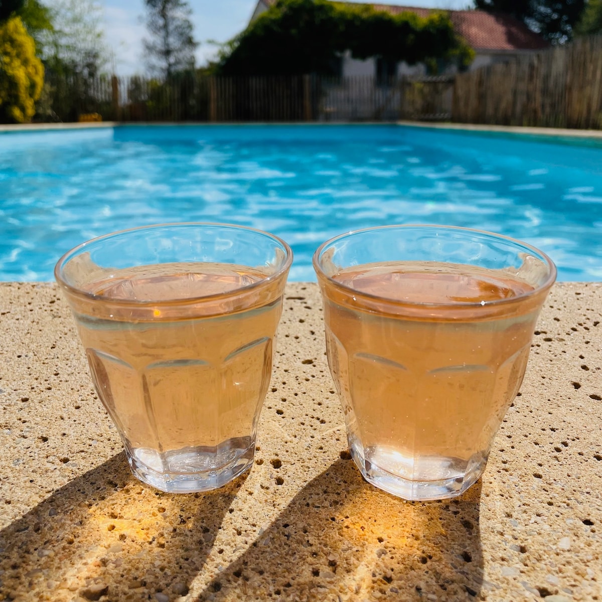 Two clear glasses are filled with a light-colored beverage, placed on a textured stone edge. The inviting blue water of a swimming pool is visible in the background, surrounded by a wooden fence and greenery under a bright sky.