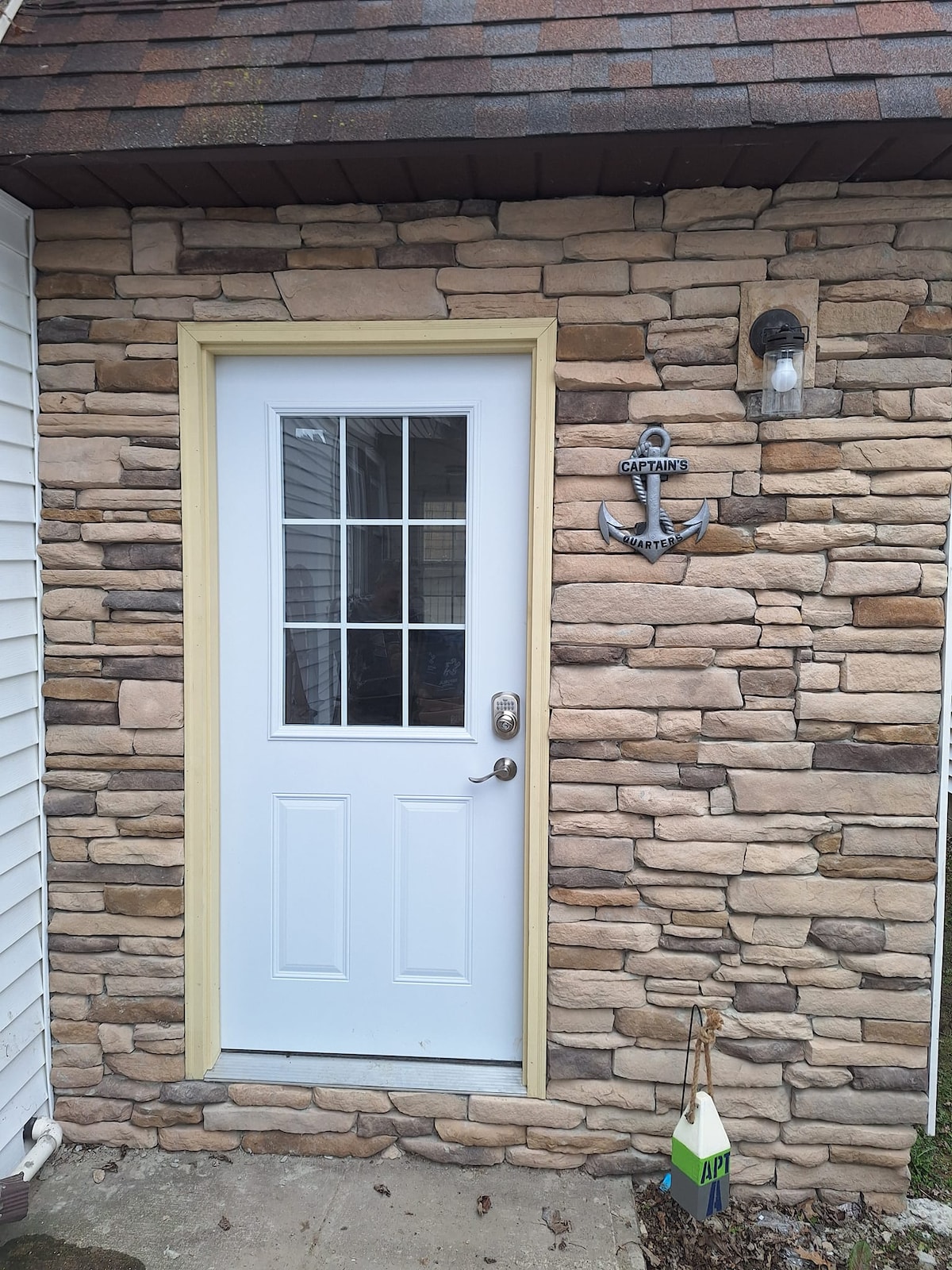 The entrance features a white door framed by decorative stone walls, accented with a nautical anchor detail. A small glass window above the door allows natural light to filter in, while a wall-mounted light fixture offers illumination.