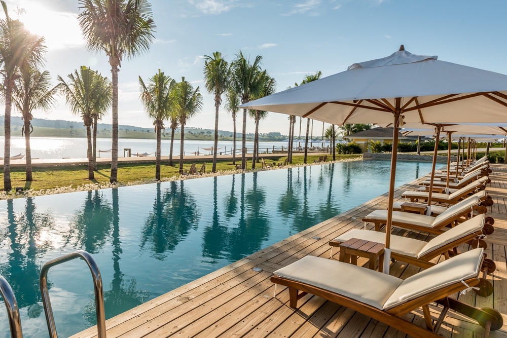 A serene pool area is framed by palm trees, featuring wooden lounge chairs with umbrellas. The inviting pool reflects the clear sky and borders a scenic view of the water, enhancing the outdoor relaxation space.