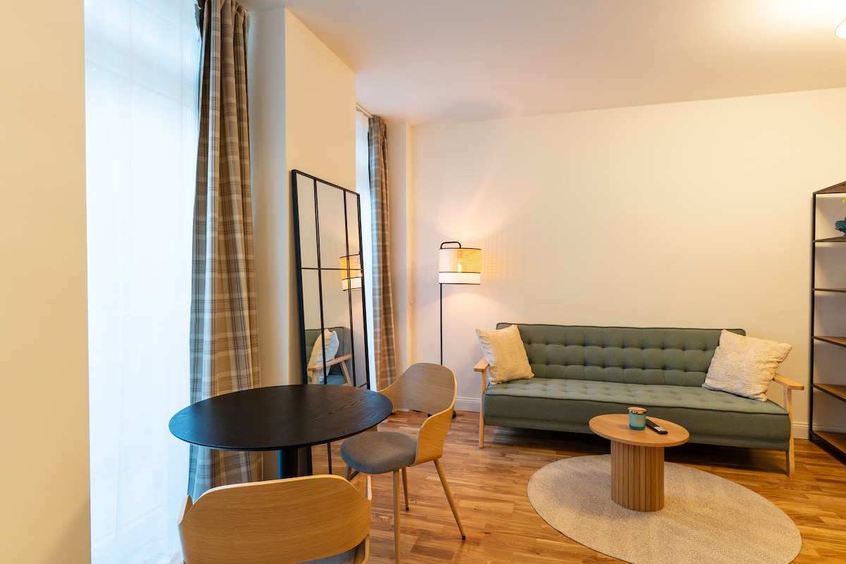 A cozy living space features a green sofa positioned against a light wall, accented by a floor lamp and a mirror. A round wooden table with two chairs invites casual dining, while warm wooden flooring adds to the room's overall comfort.